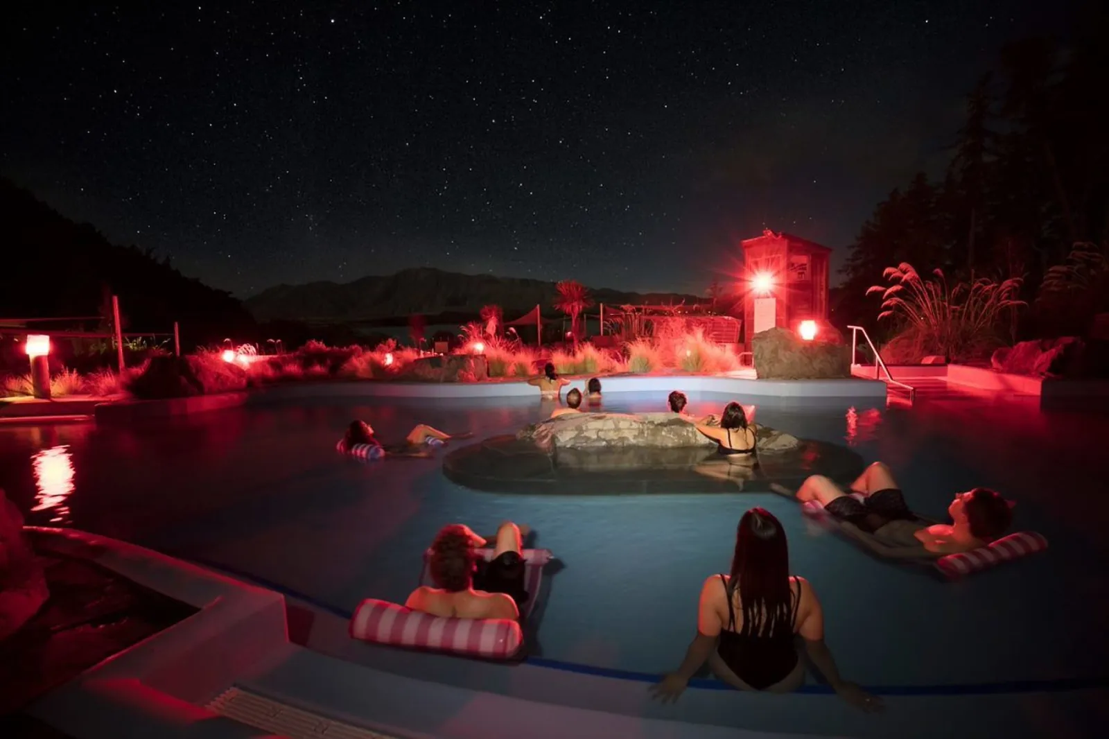 Hot pools glowing red at night with guests watching the sky