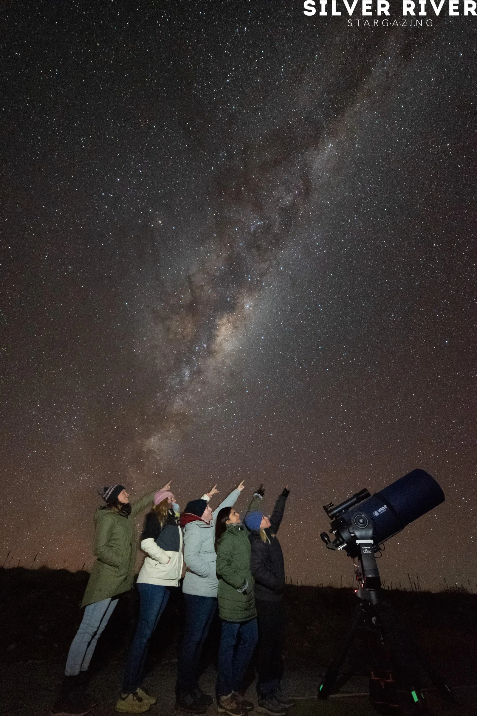 Guests raising their arms under the stars beside a large telescope